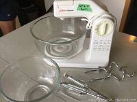 Overall view of the white Sunbeam Mixmaster mixer with two glass mixing bowls and metal attachments on a white surface.