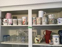 Four shelves filled with assorted mugs on top two shelves, clear glassware on lower left, and cream/sugar bowls on lower right.