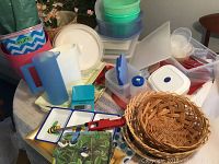 Photo showing assorted plastic food storage containers, ceramic serving trays with bird motifs, woven baskets, blue pitcher, barbecue lighter, and decorative waste baskets on a round table.