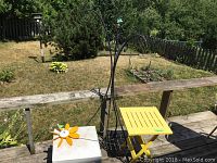 Photo showing the three items on a deck rail: black iron tripod shepherd hook, yellow plastic collapsible table, white plastic side table with sunflower windmill on top.