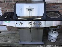 Front view of Blue Ember stainless steel propane barbecue grill with four knobs, side burner on right, cooking utensils on left side shelf, and propane tank underneath right shelf.