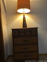 Tallboy dresser with retro ceramic lamp on top, lamp is switched on showing fabric shade illuminated.