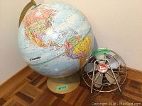 The 16 inch Transworld spinning globe and 8 inch Torcan metal fan placed side by side on a wooden floor, showing both items and their relative sizes.