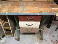 Front view of wood top workbench with paint marks and metal legs, showing three drawers in brown, white and red with metal handles.