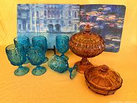 group shot of six teal goblets, two amber covered dishes and two of the placemats with artwork
