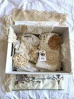 Box containing numerous cream crochet doilies and lace pieces with embroidered linen beneath