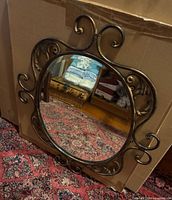 Ornate brass-tone scrollwork wall mirror leaning against cardboard backdrop