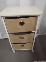 Front view showing three beige fabric drawers with grommet pulls in white metal frame on casters