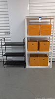 black metal 3-tier shelf beside white plastic 4-tier shelf with four orange fabric bins