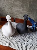 Two ceramic bird bisques (white and black) beside blue gargoyle on lace mat