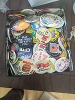 Open container filled with numerous round pinback buttons showing varied graphics and slogans