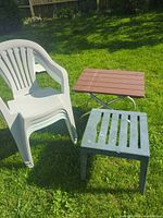 White stackable chair(s), gray side table, brown slatted folding table on grass