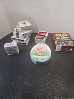 Group shot showing all items: UNO No Mercy box, iota tin, Little Eggs tin, loose UNO deck, stack of standard playing card decks