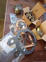 Overview of two carburetor bodies, loose gaskets, boxed items on tabletop