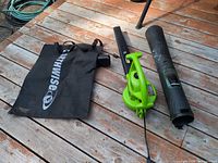 Green Earthwise corded electric leaf blower/vacuum with power cord, black collection bag, two black tube attachments on wooden deck