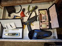 Drawer view showing glue gun, infrared thermometer, tachometer in case, hand-drill set, pliers, orange drill adapter