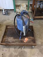 Front view of blue steel sandblaster on wooden dolly with hose and nozzle