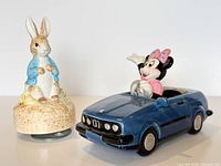 Front view of both ceramic music boxes – Peter Rabbit on straw base and Minnie Mouse driving blue convertible
