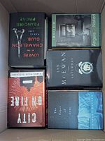 Top layer of six hardcover novels inside box (e.g., Lovers at the Chameleon Club, City on Fire, Saturday)