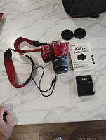 Red Canon DSLR with 18-55mm lens, strap, charger, lens caps, manual, bag partially visible