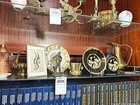 Shelf showing copper pitcher, brass bowl, framed print, porcelain plate, bronze figurine, brass cup, black lacquered items, brass pitcher