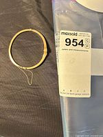 Overall view of gold hinged bangle bracelet beside lot tag, showing oval shape and attached safety chain