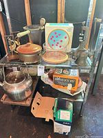Full lot on shelving showing boxed Römertopf, kettle, press, bookends, strainer, mortar, saucepan, boxed items