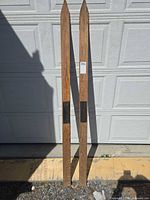 Full view of both wooden skis leaning against garage door showing length, natural wood finish, black footrest pads, pointed tips