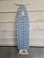 ironing board standing folded showing patterned blue cover and white frame