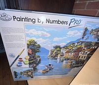 Front view of boxed Painting by Numbers Pro kit showing harbour scene artwork and included materials