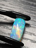 Rectangular jelly opal held in tweezers showing green, blue and orange pinfire