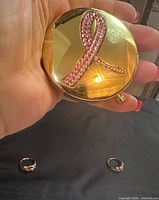 Gold-tone compact with pink crystal ribbon held in hand, rings visible below
