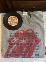 Front of grey Rolling Stones tee with tongue logo, lyrics, and vinyl record placed on shirt