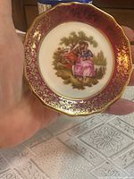 Front view of miniature Limoges plate showing burgundy rim, gold filigree, courting couple motif
