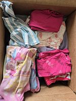 Top-down view of open box showing assorted size 4T girls garments including FILA T-shirt, striped dress, printed tops and fleece towel