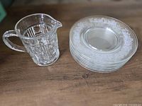 Clear glass cream pitcher beside stack of five etched dessert plates
