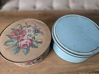 Beige floral tin and blue tin side by side on tabletop showing lids and wear