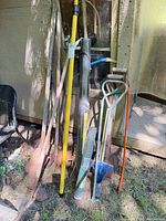 Group of garden tools leaning against wall including shovels, post hole digger, fencing roll