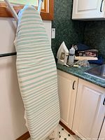 Full-size ironing board with light blue/aqua striped cover standing beside counter