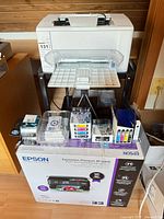 White HP printer on stand above Epson XP-6000 boxed unit with assorted ink cartridges and accessories on top