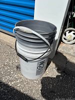 Stacked white and grey buckets with siphon hose coiled on top, showing handles and caution label