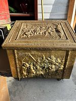 Front and top view of embossed metal coal box showing decorative scenes and casters