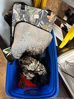 Blue tote holding two camouflage seat cushions, rolled camo fabric, and additional hunting clothing