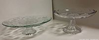 Two clear pressed glass pedestal cake stands, floral flat plate and scalloped rim styles