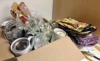 Open box showing mugs, glassware, rolling pin, wooden spoons, citrus juicer, textile stacks
