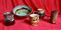 Five handmade glazed ceramic pieces on red backdrop: bowl, pitcher, three mugs