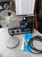 Front and top view of projector with attached clear reel, reel-can lid, power cord and manual visible