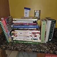 Full lot showing stacked hardcovers, spiral and comb-bound cookbooks on countertop