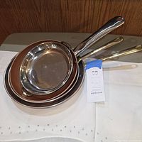 Three nesting copper-exterior frying pans with brown ceramic interiors and long handles