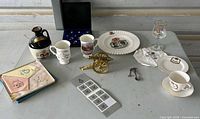 Overview of lot showing plate, mugs, jug, spoons case, brass cannon, napkins, glass and other items on table